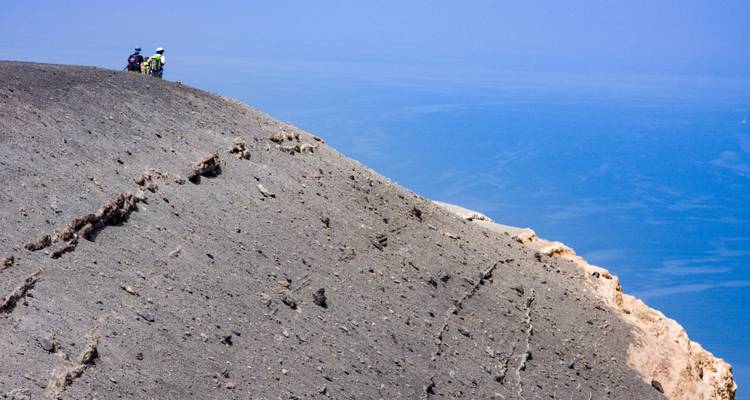 Eine weite vulkanische Landschaft mit Blick auf das Meer und Wanderern auf dem Gipfel.