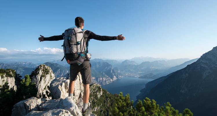 Rückansicht eines Rucksacktouristen, der triumphierend auf einem felsigen Grat steht und auf den Gardasee und die entfernten Alpen blickt.