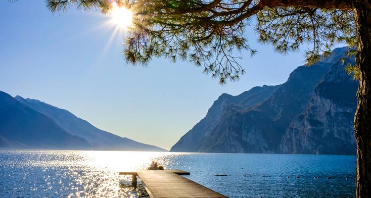 Ein einzelner Reisender sitzt am Ende eines hölzernen Stegs und blickt auf einen glitzernden Alpensee mit Sonnenlichteffekt.