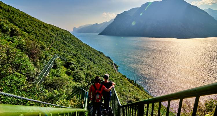 Ein Paar macht eine Pause auf einem malerischen Wanderweg hoch über dem Gardasee und blickt auf das glitzernde Wasser und die Berge.