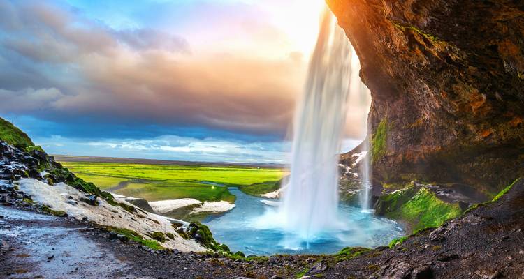 Der Seljalandsfoss-Wasserfall stürzt von einer moosbedeckten Höhlenkante in ein leuchtendes grünes Tal bei Sonnenuntergang.