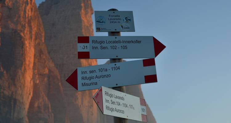 Wanderwegweiser, die auf verschiedene Rifugios hinweisen und von hoch aufragenden orangefarbenen Dolomitenwänden bei Sonnenaufgang eingefasst sind.