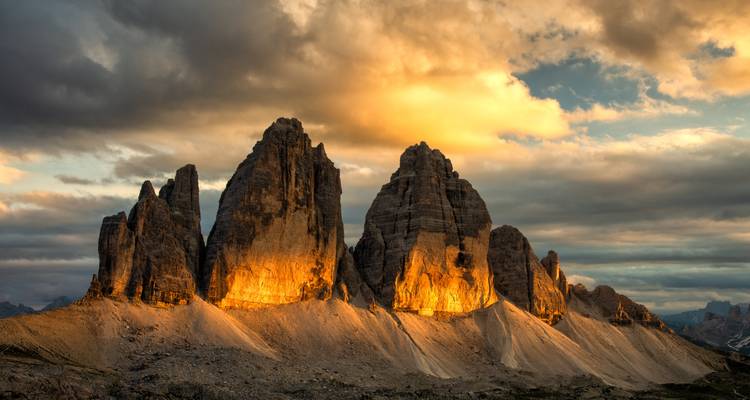 Die gezackten Tre-Cime-Gipfel leuchten bei einem dramatischen Sonnenuntergangshimmel voller düsterer Wolken in Orange auf.