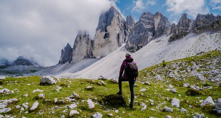 Ein einsamer Wanderer mit einem Rucksack steht auf einem grasbewachsenen Hang und blickt auf die nebelumhüllten Felsspitzen der Tre Cime.