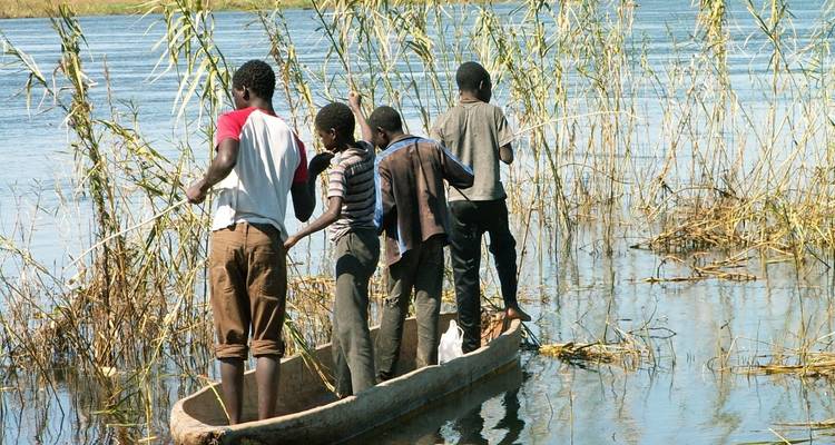 Vier einheimische Jungen, die in einem ausgehöhlten Kanu zwischen hohen Schilfrohren auf einem ruhigen Fluss balancieren.