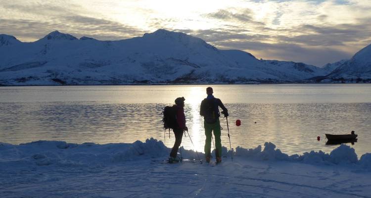Menschen, die Ski fahren an einem Fjord mit schneebedeckten Bergen im Hintergrund.