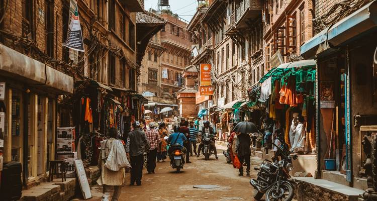 Drukke straatscène met mensen, motorfietsen en winkels langs een smalle steeg.