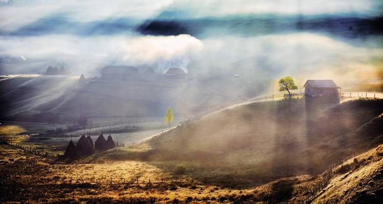 Mistig landelijk landschap met zonnestralen en kleine huizen.