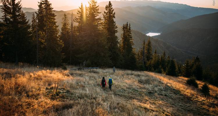Twee mensen die naar een ondergaande zon lopen met bergen op de achtergrond.