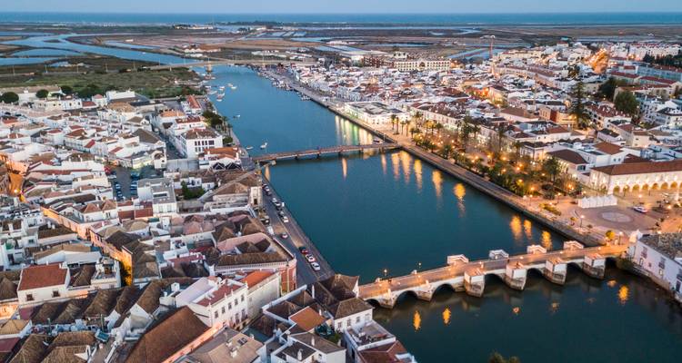 Evening aerial of Tavira with illuminated bridge and salt marshes fading into dusk.