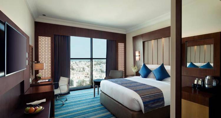 Habitación de hotel con una cama grande, cojines azules y vista al exterior.