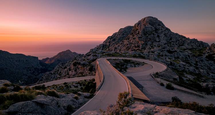 Serpentinenstraße in den Bergen schlängelt sich einer felsigen Bergspitze unter einem Himmel in Pastelltönen bei Sonnenuntergang an der Küste von Mallorca entgegen.