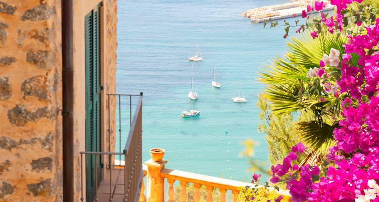 Lebendige rosa Bougainvilleen rahmen eine türkisfarbene Bucht mit kleinen Segelbooten unter einer sonnenbeschienenen Terrasse ein.