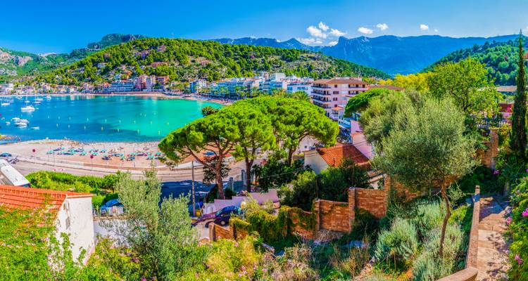 Panoramablick auf eine lebhafte mediterraneische Strandstadt mit smaragdgrüner Bucht und den fernen Gipfeln der Serra de Tramuntana.