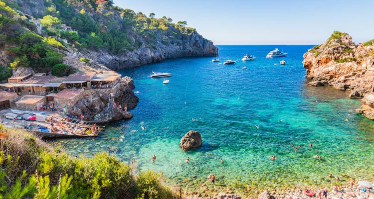 Kristallklare Bucht mit Badegästen, Yachten und Felsenriffen, die zwischen steilen Klippen auf Mallorca eingebettet sind.