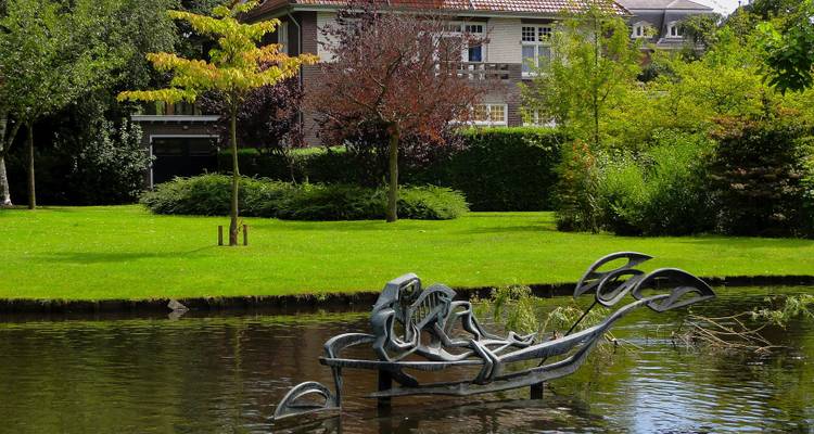 Modern abstract metal sculpture afloat on a tranquil pond in a leafy urban park.