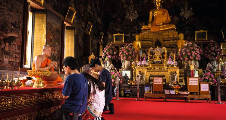 Des fidèles agenouillés devant un moine à l'intérieur d'un temple somptueux rempli de statues de Bouddha dorées et d'offrandes.