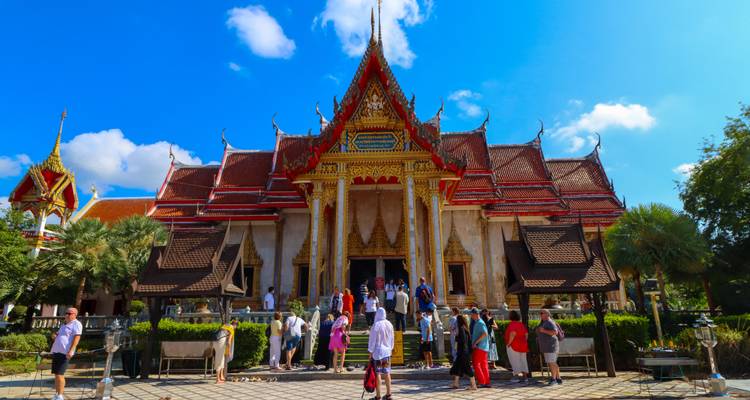 Les visiteurs entrant dans un temple orné de Phuket avec un toit rouge et des décorations dorées sous un ciel dégagé.
