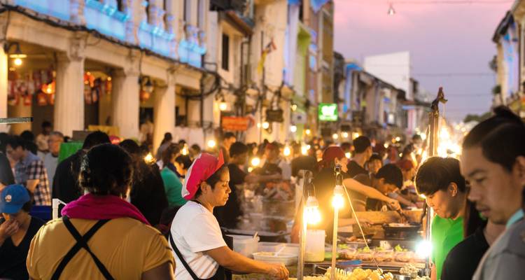 Marché de rue animé en soirée avec des étals alimentaires éclairés par des ampoules brillantes et des visiteurs qui font leurs courses.