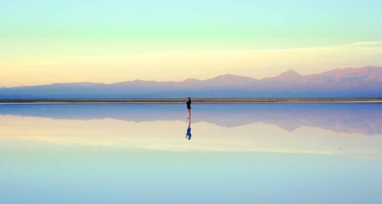 Eine einzelne Person, die bei Dämmerung über eine ruhige Salzebene mit Bergen wandelt.
