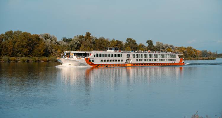 Large river cruise ship navigating through a calm river with trees in the background.