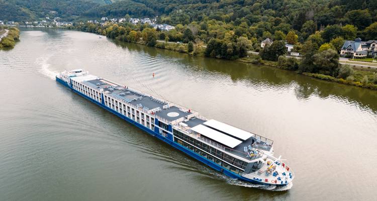 A modern blue-and-white river cruise ship sails through a tranquil green valley river.