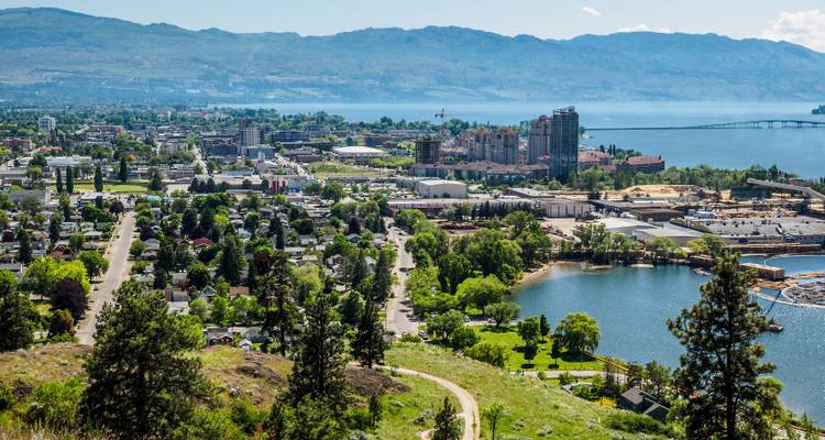 Aerial view of a city with a lake, forested areas, and distant mountains, likely Kelowna, Canada.