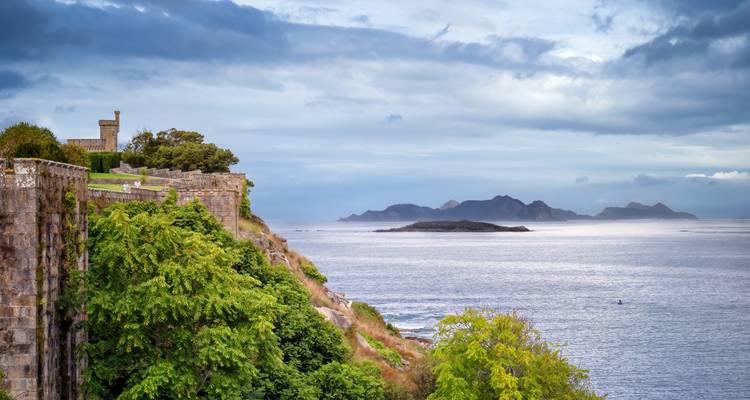 Fortaleza de piedra en un promontorio frondoso que domina una tranquila bahía del Atlántico con las Islas Cíes a lo lejos.