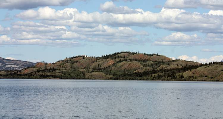 Calm lake with low wooded hills and scattered clouds on a bright afternoon.