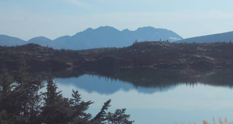 Still lake reflecting low jagged hills under a hazy pale blue sky.