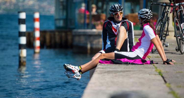 Cyclists resting by the water with bicycles and mountains in the background.