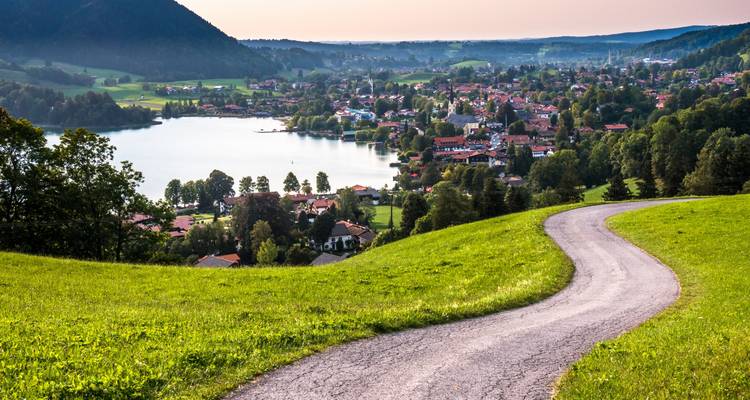 Winding road leading down to a lakeside town with mountains in the background.