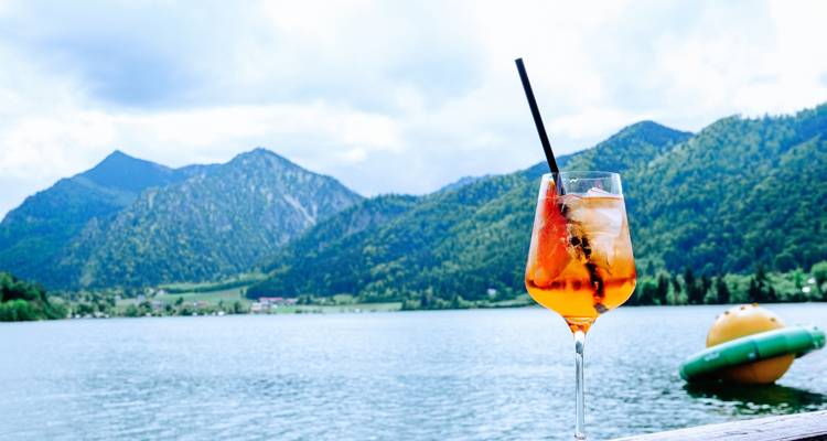 Glass of Aperol Spritz with mountains and a lake in the background.