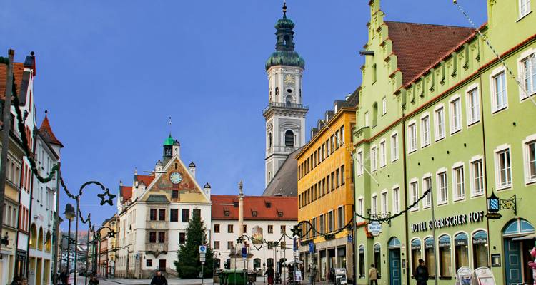 Bunte Fassaden säumen eine Fußgängerstraße in einer bayerischen Stadt, die zu einem hohen Kirchturm unter einem klaren blauen Himmel führt.