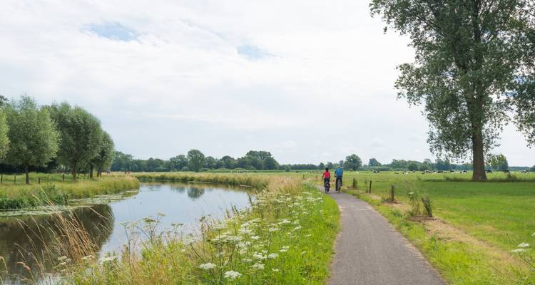 Zwei Radfahrer fahren auf einem ruhigen gepflasterten Weg neben einem stillen Kanal, der von flachen grünen Wiesen umgeben ist.