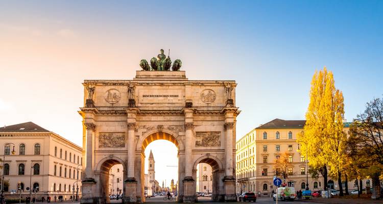 Der Siegestor in München erstrahlt im sanften Morgenlicht, eingerahmt von herbstlichen Bäumen auf beiden Seiten einer breiten Allee.