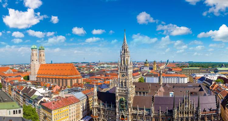 Panoramablick über Münchens Altstadt mit den Türmen der Frauenkirche und dem prunkvollen Neuen Rathaus vor einem leuchtend blauen Himmel.