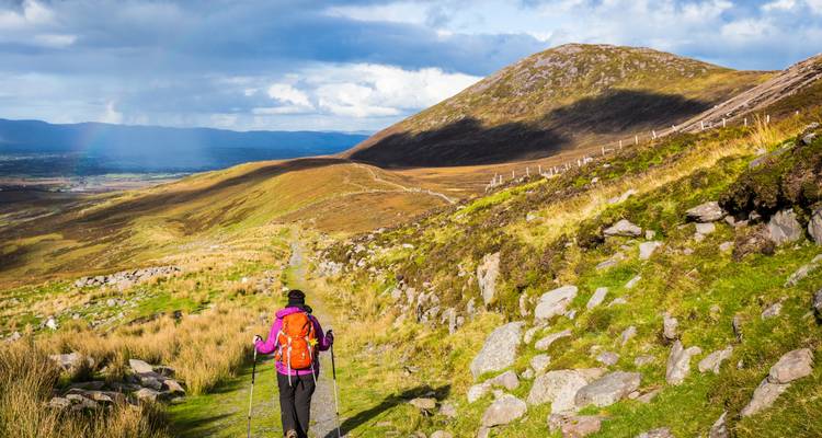 Een backpacker wandelt over een groen bergpad onder verspreide wolken en een mozaïek van zonlicht.