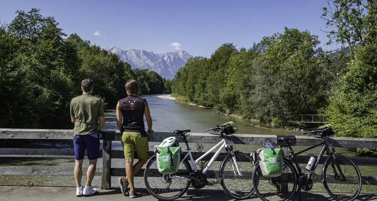 Zwei Personen mit Fahrrädern blicken auf einen Fluss und Berge.