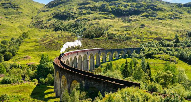 Eine Dampflokomotive überquert das imposante Glenfinnan-Viadukt inmitten üppig grüner Hügel und Wälder.