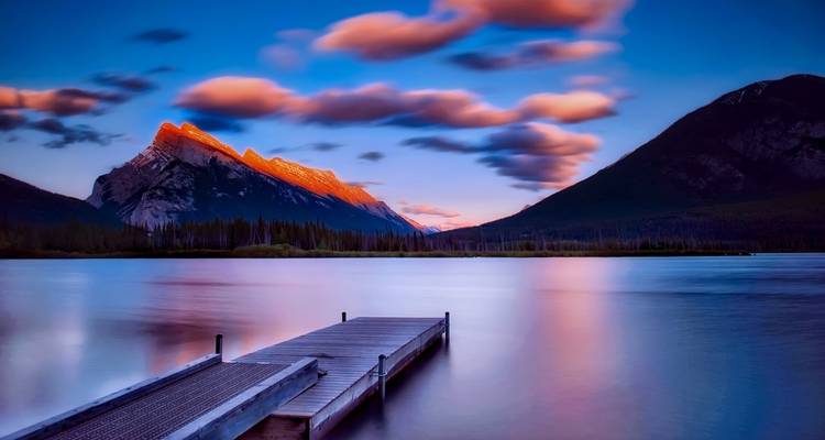 Scenic lake with a dock and dramatic mountain peaks under colorful sky.