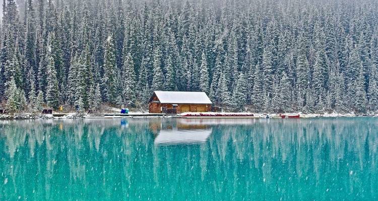 Blokhut aan een besneeuwde oever van een meer met reflecties in het water.