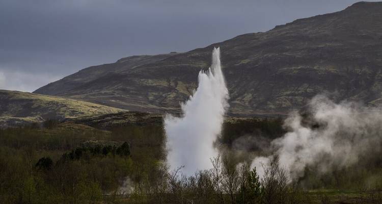 A powerful geyser erupts, sending steam and water skyward against a moody volcanic landscape.