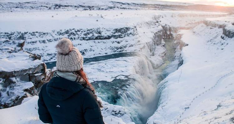 A bundled traveller looks over the snow-covered Gullfoss canyon glowing in winter sunrise light.