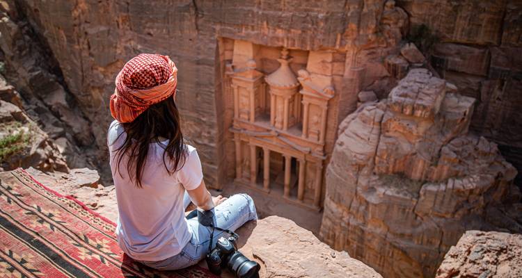 Female traveller sits on a rug at a high cliff viewpoint overlooking Petra’s Treasury far below.