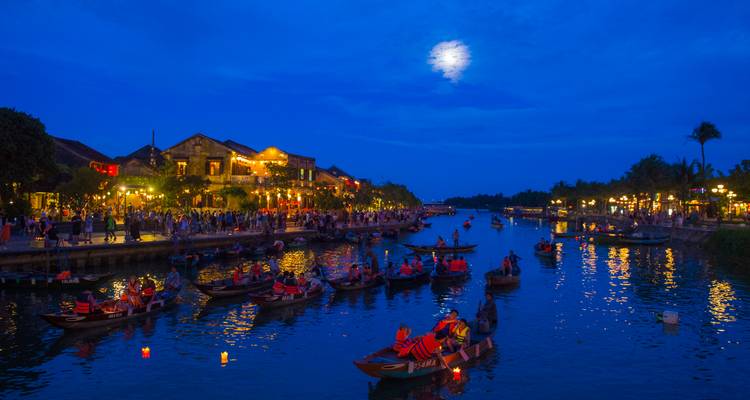 La rivière d'Hoi An éclairée par les lanternes la nuit avec des bateaux en bois transportant des visiteurs sous une lune brillante.