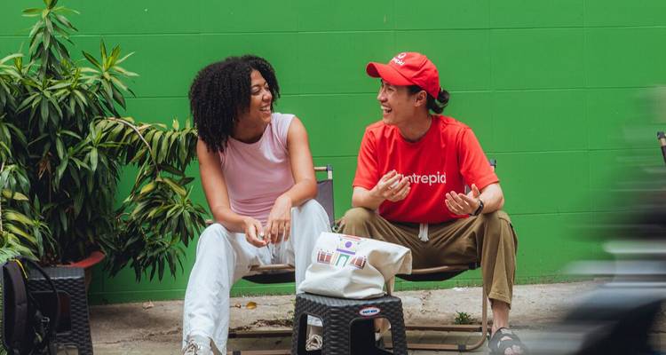 Two friends sit on low chairs chatting in front of a bright green wall on a city street.
