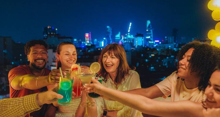 Group of travellers toast colourful cocktails on a rooftop with neon city skyline behind.