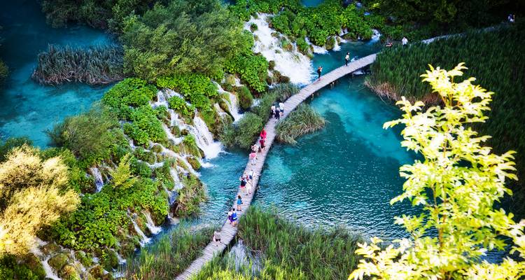 Luftaufnahme eines hölzernen Bohlenwegs, der sich über türkisfarbene Becken und kaskadierende Wasserfälle im Nationalpark Plitvicer Seen schlängelt.