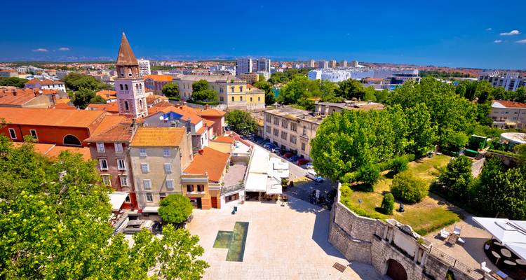 Sunny aerial of Zadar old town with bell tower, terracotta roofs and deep blue Adriatic coastline.